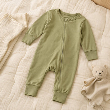 Pyjama bébé à fermeture magnétique vert olive posé à plat sur une courtepointe en coton, avec un doudou ours