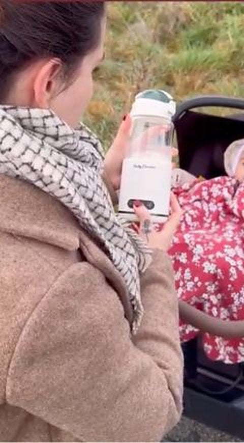 Une maman en manteau tient le chauffe-biberon Babycozidoo à côté de la poussette, en extérieur
