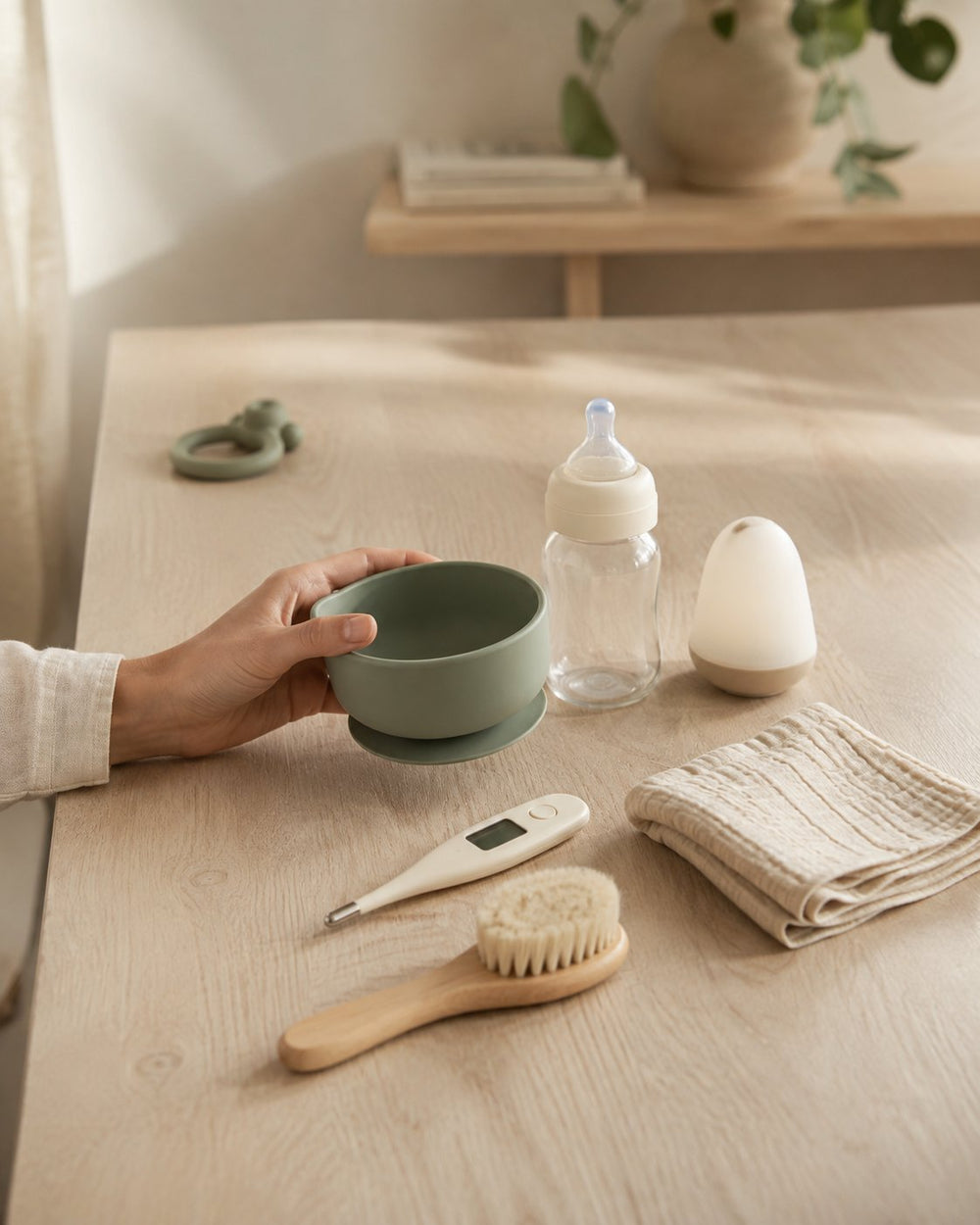Main tenant un bol en silicone vert à côté d'un biberon, d'un thermomètre, d'une brosse douce et d'un lange sur une table en bois : la sélection de produits bébé BestMomShop