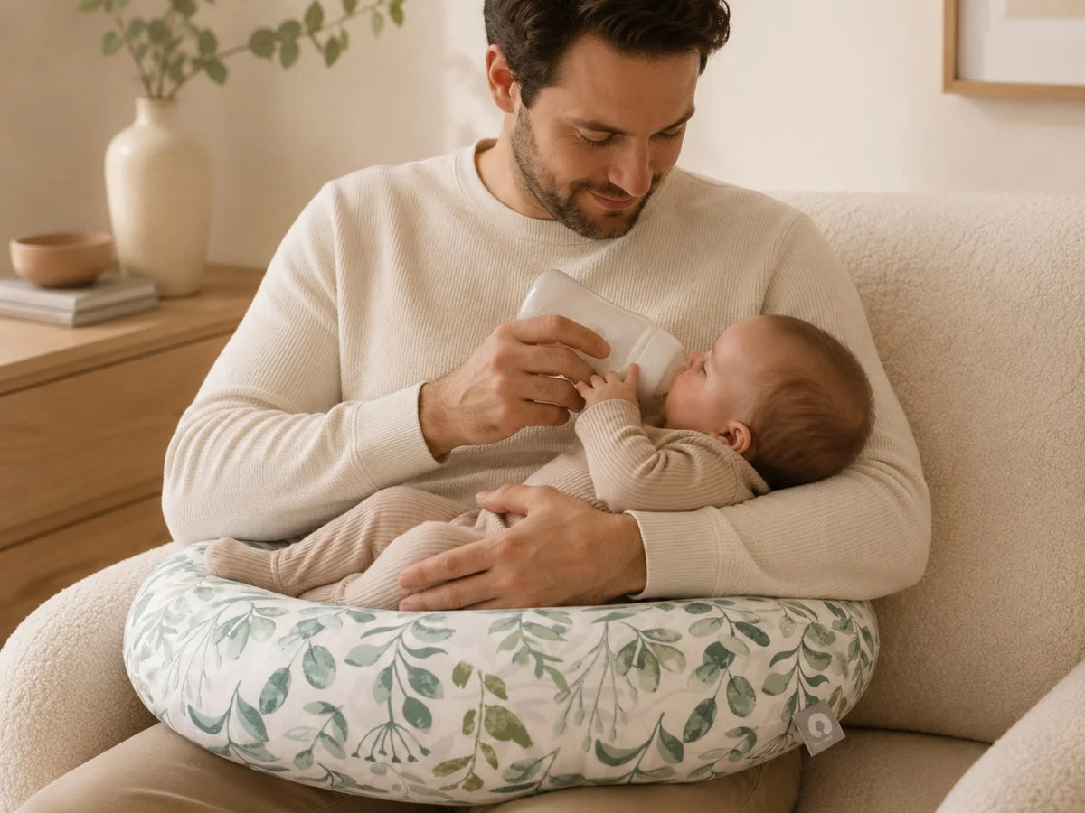 Un papa installé dans un fauteuil clair donne le biberon à son nouveau-né posé sur un coussin d'allaitement