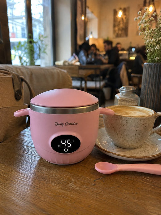 Un Babycozidoo rose posé sur la table d'un café, à côté d'un cappuccino et d'une cuillère de bébé